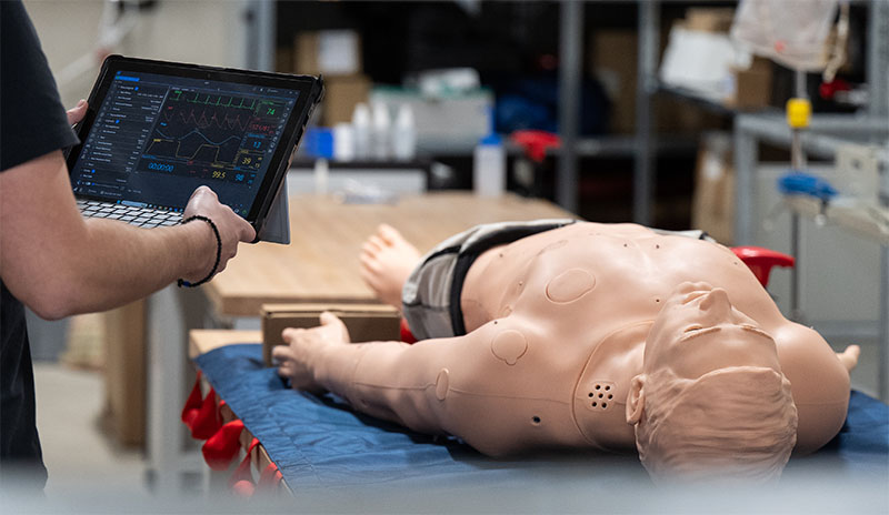 Eine Person steht mit Laptop in der Hand vor einer Simulationspuppe bei Mefina Medical