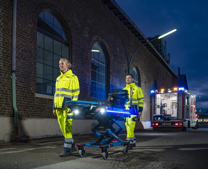 Szenenbild_Stollenwerk_Tragesystem Trage von Stollenwerk auf Asphalt mit 2 Rettungssanitätern, welche sie zur Kamera hin schieben