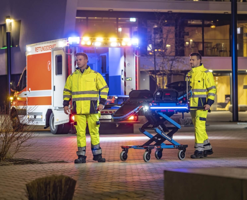 Stollenwerk_System_Szenenbild Fahrtrage vom Rettungsdienst mit 2 Sanitätern um diese rum und offenen Rettungswagen + Gebäude im Hintergrund