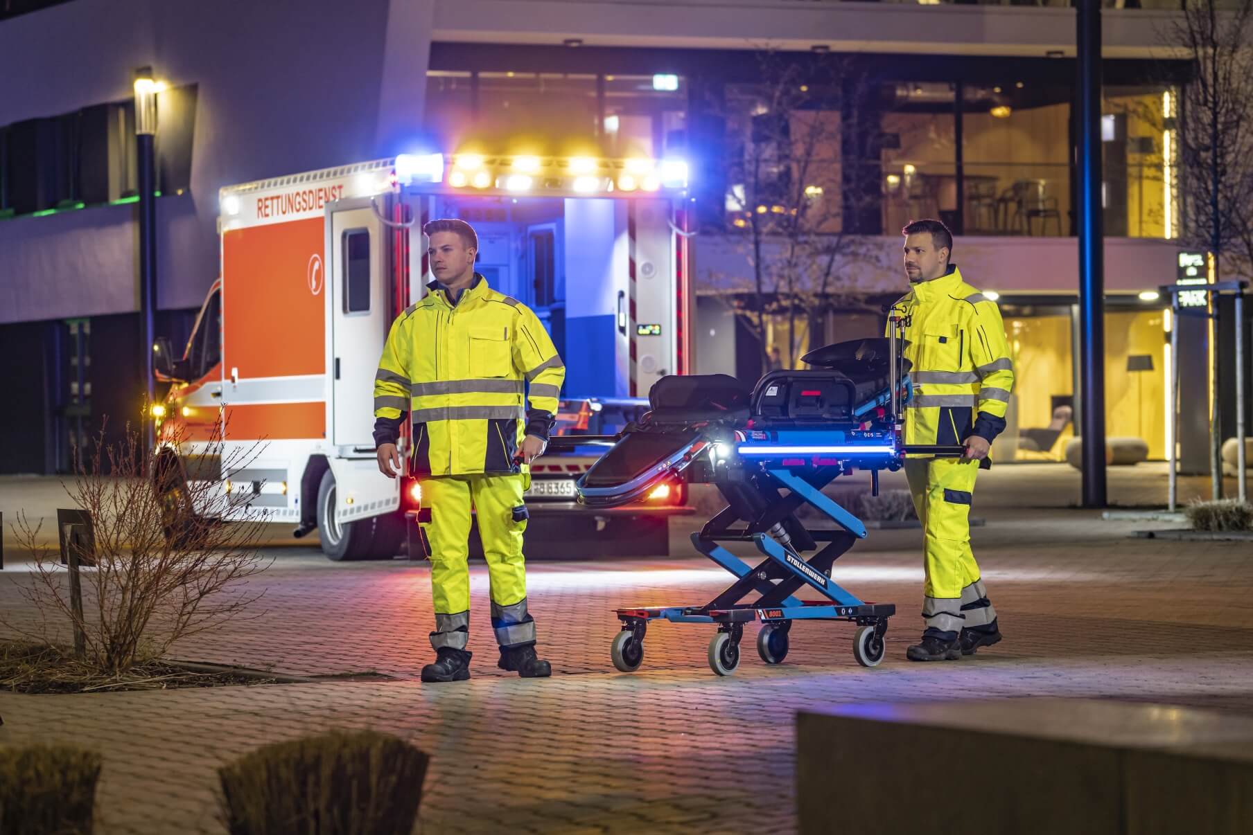 Stollenwerk_System_Szenenbild Fahrtrage vom Rettungsdienst mit 2 Sanitätern um diese rum und offenen Rettungswagen + Gebäude im Hintergrund